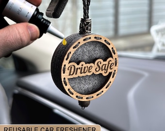 Puede incluir: Un ambientador de coche redondo con las palabras "Drive Safe" recortadas en la madera. El ambientador está hecho de madera y fieltro gris. Se muestra una mano aplicando un líquido de una botella pequeña al ambientador. Las palabras "REUSABLE CAR FRESHENER" están en la parte inferior.