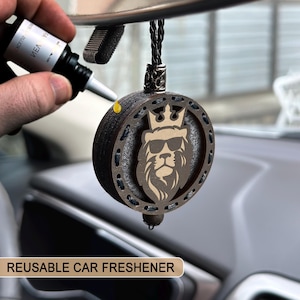 Puede incluir: Un ambientador de coche reutilizable con un león con gafas de sol y una corona. El disco de madera marrón tiene un borde decorativo y un cordón negro. El texto "REUSABLE CAR FRESHENER" está en la parte inferior.