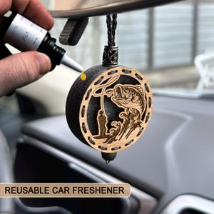 Könnte beinhalten: Ein runder Auto-Lufterfrischer aus Holz mit einem Angelmotiv. Das Design zeigt einen Fisch, der aus dem Wasser springt, mit einem Angler. Der Text "REUSABLE CAR FRESHENER" steht unten. Der Lufterfrischer hängt an einer schwarzen Schnur.