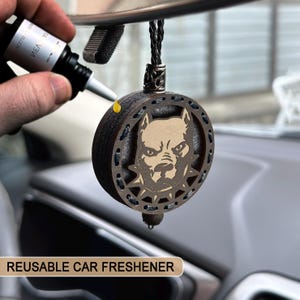 Puede incluir: Un ambientador de coche circular marrón oscuro con un diseño de pitbull. El ambientador se está rellenando con una botella negra. El texto "REUSABLE CAR FRESHENER" está en la parte inferior de la imagen.