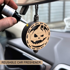 Pode incluir: Um ambientador de carro reutilizável em forma de abóbora de madeira com uma borda preta e um rosto de jack-o'-lantern esculpido. O ambientador pende de um cordão preto e está a ser reabastecido. O texto "REUSABLE CAR FRESHENER" está na parte inferior.