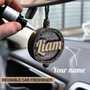 May include: A personalised car air freshener with the name "Liam" in a wooden design. The reusable freshener is round with a dark brown frame and a grey interior. A hand is shown adding fragrance from a small bottle. The text "REUSABLE CAR FRESHENER" is at the bottom.