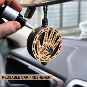Peut inclure: Un désodorisant de voiture rond en bois avec les mots "Dad!" et "Super Dad" gravés. Un flacon noir est utilisé pour parfumer le désodorisant. Le texte "REUSABLE CAR FRESHENER" est en bas.