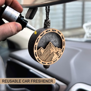 Può includere: Un profumatore per auto rotondo in legno con un design a montagna. Il profumatore ha una cornice in legno scuro con una scena di montagna in legno chiaro al centro. Il testo "REUSABLE CAR FRESHENER" è in basso.
