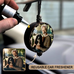 May include: A black, round reusable car air freshener with a photo of a man and a dog. The freshener is hanging from a braided black cord. A hand is adding a yellow liquid to the freshener. The text "REUSABLE CAR FRESHENER" is visible.