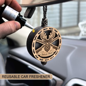 May include: A round, wooden car air freshener with a deer and crossed rifles design, and the text "Born to Hunt". The freshener is black and light brown, with a black cord for hanging. A bottle of scent is being applied to the freshener.