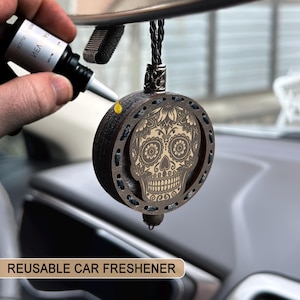 Puede incluir: Un ambientador de coche reutilizable con un diseño de calavera tallada. El ambientador redondo de madera marrón oscuro cuelga de un cordón negro. La calavera tiene elementos florales y decorativos. El texto "REUSABLE CAR FRESHENER" está en la parte inferior.