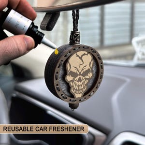 May include: A reusable car freshener with a skull design. The round, wooden freshener features a detailed skull graphic in the center, surrounded by a decorative border. The freshener hangs from a black cord and is being refilled with a liquid.