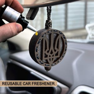May include: A round, wooden car air freshener with a Ukrainian trident emblem. The freshener is dark brown with a silver-colored border and a black cord for hanging. A small bottle is dispensing liquid onto the freshener. The text "REUSABLE CAR FRESHENER" is at the bottom.