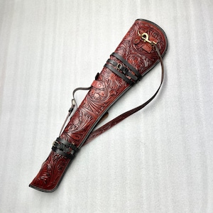 May include: A reddish-brown leather quiver with detailed floral tooling. It has black leather straps with buckles, a shoulder strap, and a brass-coloured clasp. The design features embossed patterns, suitable for archery. The quiver is approximately 75 cm in length.