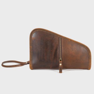 Könnte beinhalten: Eine dunkelbraune Ledertasche mit asymmetrischer, geschwungener Form und glatter, matter Oberfläche. Sie verfügt über ein vertikales Reißverschlussfach mit passendem Lederzugband und eine robuste Handschlaufe aus Leder, die seitlich mit einer Niete befestigt ist.