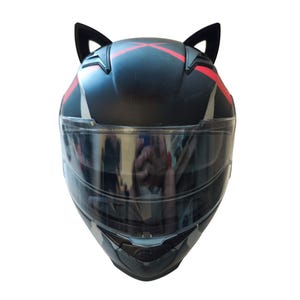Może przedstawiać: Czarny kask motocyklowy z przezroczystym wizjerem i czerwonymi akcentami. Kask ma dwa czarne kocie uszy przymocowane do g&oacute;ry.
