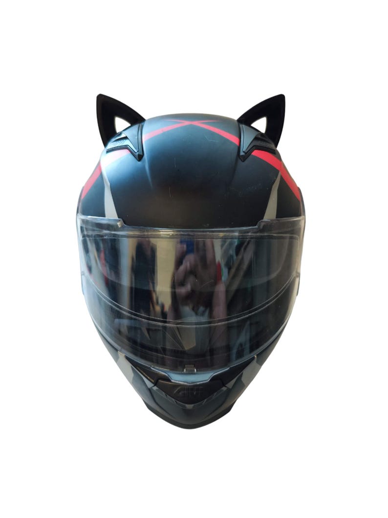 Może przedstawiać: Czarny kask motocyklowy z przezroczystym wizjerem i czerwonymi akcentami. Kask ma dwa czarne kocie uszy przymocowane do g&oacute;ry.