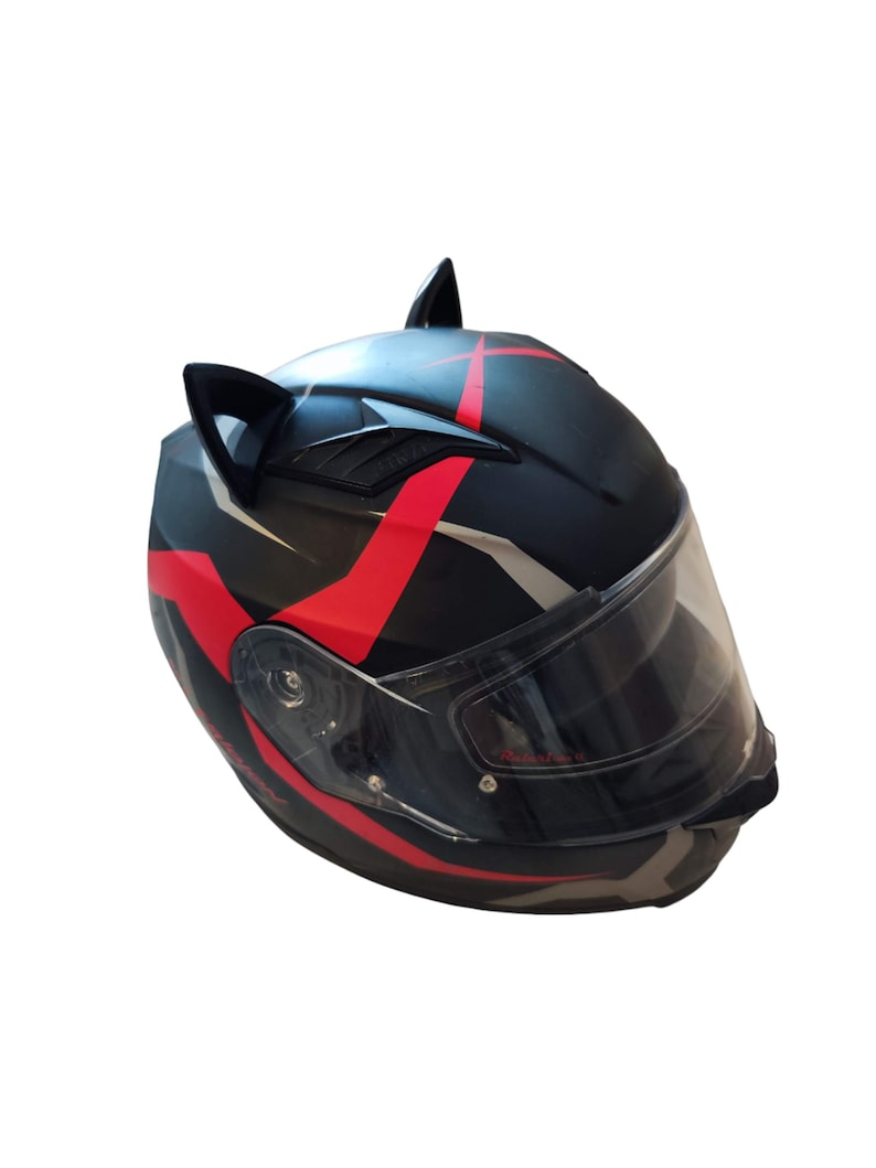Może przedstawiać: Czarny kask motocyklowy z czerwonymi i białymi akcentami i czarnymi uszami kota. Kask ma przezroczysty wizjer.
