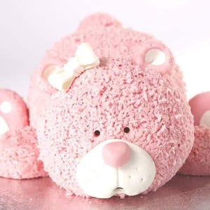 Puede incluir: Un pastel de oso de peluche rosa con glaseado texturizado. El oso tiene detalles blancos en la cara y las patas, un lazo blanco y una nariz rosa. El pastel está sobre una superficie plateada.