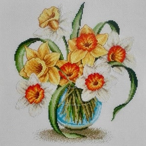 Può includere: Opera d'arte a punto croce di un bouquet di narcisi in un vaso blu. I fiori sono gialli e bianchi con centri arancioni, circondati da foglie verdi. Il vaso poggia su una superficie neutra, creando un'esposizione floreale vibrante.