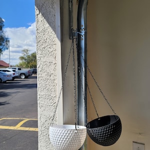 May include: Two hanging planters, one white and one black, with a honeycomb pattern. The planters are hanging from a chain attached to a hook on a garage door.