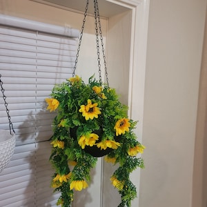 Pode incluir: Um vaso pendurado com uma corrente preta e um vaso preto cheio de uma planta verde frondosa com flores amarelas. A planta está pendurada na frente de uma janela branca com persianas.