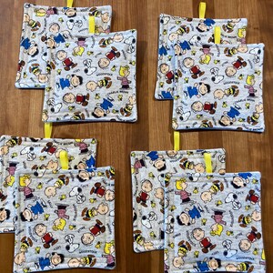Może przedstawiać: Zestaw ośmiu kwadratowych podkładek pod garnki z szarym tłem i powtarzającym się wzorem Snoopy'ego, Charliego Browna, Woodstocka i innych postaci z Peanuts. Każda podkładka ma żółtą pętlę do zawieszenia.