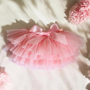 May include: A layered pink tulle skirt with a large satin bow at the waist. The skirt is designed with multiple layers of soft, sheer fabric, creating a full, fluffy appearance. The bow adds a decorative touch to the skirt.