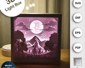 Puede incluir: Una caja de luz 3D con una luna, montañas y árboles. La caja de luz está iluminada y la luna brilla. La caja de luz está hecha de papel y es un proyecto artesanal. La imagen también incluye el texto "3D Light Box", "SVG", "DXF", "EPS", "PDF", "Enjoy Your Shadow Box", y "+ Free Frame Box".