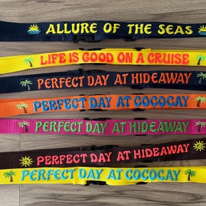 May include: Seven nylon webbing straps with black plastic side-release buckles. The straps feature embroidered text and icons: ALLURE OF THE SEAS, LIFE IS GOOD ON A CRUISE, and PERFECT DAY AT HIDEAWAY or COCOCAY, accented with palm tree and sun motifs.