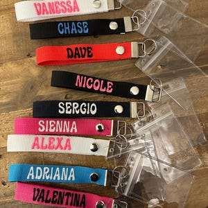 Puede incluir: Una colección de llaveros personalizados en varios colores, cada uno bordado con un nombre en un color contrastante. Los llaveros incluyen nombres como "Vanessa", "Chase" y "Dave". También se ven porta-identificaciones de plástico transparente.