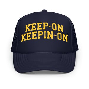 May include: Navy blue trucker hat with the text "KEEP ON KEEPIN ON" in yellow. The hat has a mesh back and a curved brim. The text includes star accents.