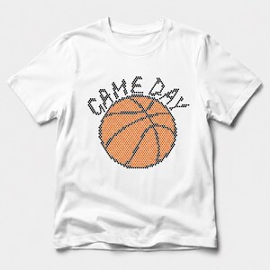 May include: A white short-sleeved t-shirt featuring a graphic design on the chest. The design consists of a basketball rendered in orange and black dots, with the text "GAME DAY" arched above it in black dots, creating a textured appearance.