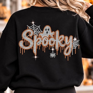 Op de afbeelding: Een zwart sweatshirt met lange mouwen en een strass-afbeelding op de achterkant. Het ontwerp toont het woord Spooky in oranje en witte kristallen met een druipend effect, vergezeld door een geest, spinnenwebben en een kleine spin.