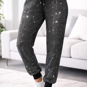 May include: Black sweatpants covered in small, sparkling embellishments. The pants have a drawstring waist and black cuffs at the ankles. The person is wearing white sneakers. The background includes a white couch and a light-colored rug.