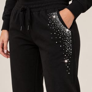 Puede incluir: Pantalones de chándal negros con cintura elástica con cordón y un bolsillo lateral adornado con un patrón en cascada de pedrería blanca de varios tamaños, creando un efecto brillante a lo largo de la cadera y el muslo.