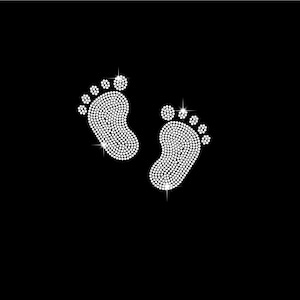 May include: Two sparkling white rhinestone baby footprints on a black background. The footprints are detailed with individual rhinestones, creating a shimmering effect. The image is simple and elegant, perfect for baby-themed crafts or designs.