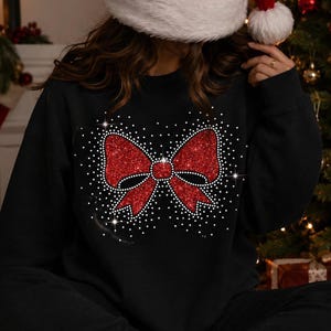 Op de afbeelding: Een zwart sweatshirt met lange mouwen en een grote, centrale afbeelding van een rode glitterstrik. De strik is omringd door een verspreiding van kleine witte stippen die een sprankelend effect nabootsen. Het sweatshirt heeft een ronde hals.