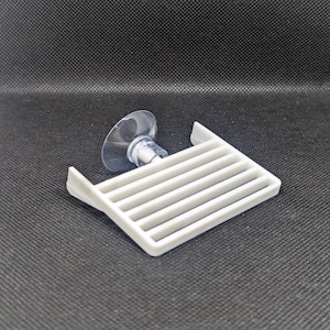 Könnte beinhalten: Weißer Plastik-Geschirrhalter mit Saugnapfbasis. Der Halter hat fünf horizontale Stäbe und ist zum Halten von Geschirr oder anderen Gegenständen gedacht.