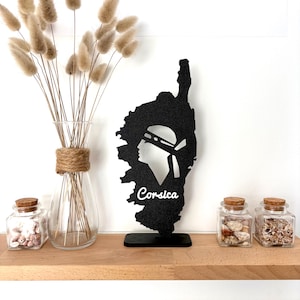 Puede incluir: Una silueta negra de la isla de Córcega con el perfil de una persona con una diadema y la palabra "Corsica" en blanco. La imagen está sobre una estantería de madera con un jarrón de flores secas y pequeños tarros de cristal con tapas de corcho.