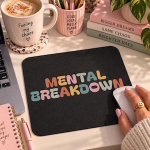 May include: A rectangular black mouse pad featuring the text MENTAL BREAKDOWN in a multi-colored, rounded, retro-style font. The letters alternate between shades of orange, pink, yellow, and teal, set against a solid black background surface.