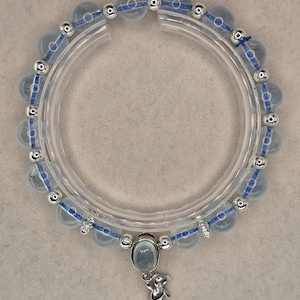 Peut inclure: Un bracelet de perles bleu clair avec des détails argentés et un petit charm en forme d'ours en peluche. Le bracelet est composé de perles rondes et translucides enfilées sur un cordon bleu, entrecoupées de perles argentées. Une pierre ovale bleu clair et un charm ours en argent pendent.