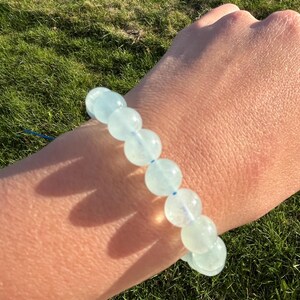 Peut inclure: Un bracelet de perles d'aigue-marine bleu clair. Le bracelet est composé de perles rondes et translucides enfilées sur un cordon bleu. Le bracelet est porté au poignet, avec de l'herbe verte en arrière-plan.