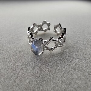 Può includere: Un anello d'argento con un design delicato e intricato che presenta un motivo a stella ripetitivo. L'anello ha una pietra preziosa ovale trasparente al centro.