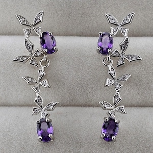 May include: A pair of silver-toned drop earrings featuring a butterfly design. Each earring has three butterfly accents with clear stone embellishments and a dangling oval-cut, deep purple gemstone. The earrings are displayed on a light gray surface.