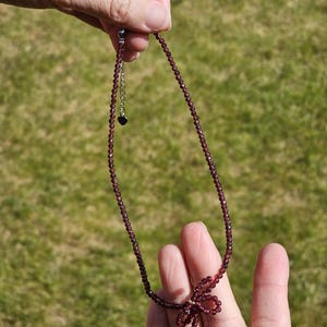 May include: A delicate necklace made of small, dark red beads with a bow at the end. The necklace has a small heart charm on the chain.