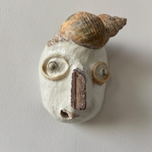 Puede incluir: Una peculiar escultura de rostro de cerámica hecha a mano con una concha marina en la cabeza. La cara es blanquecina con ojos de concha y una nariz rectangular marrón. La escultura tiene una superficie texturizada y una pequeña boca abierta.