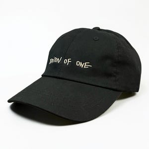 May include: A black baseball cap featuring the text "EDITION OF ONE" embroidered in white thread across the front panels. The cap has a curved brim, a structured crown with a button on top, and visible stitching details along the brim and panels.