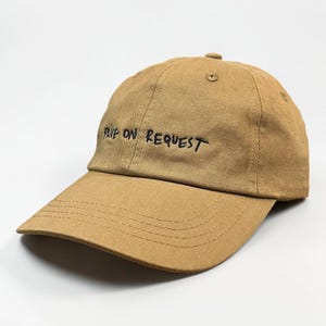 May include: A tan baseball cap featuring the embroidered black text "LIVE ON REQUEST" across the front panels. The cap has a structured crown, a curved brim with visible stitching, and a button at the top center of the fabric dome.