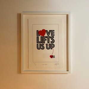Puede incluir: Una impresión enmarcada con el texto "LOVE LIFTS US UP" en letras negras. Un globo en forma de corazón rojo está unido a la palabra "LOVE" y un corazón rojo más pequeño con la palabra "LOVE" se encuentra en la parte inferior de la impresión.