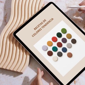 May include: A digital tablet shows the text "RECETAS DE COLORES TENDENCIA" and a selection of colourful circular objects. A wooden, wave-shaped sculpture is on the left. A hand holds a stylus, and another hand touches the tablet.
