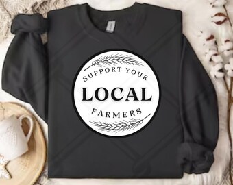 May include: Black crewneck sweatshirt with the text "SUPPORT YOUR LOCAL FARMERS" in a white circle with wheat stalks. The sweatshirt is displayed on a neutral-toned surface with a white mug and decorative elements.