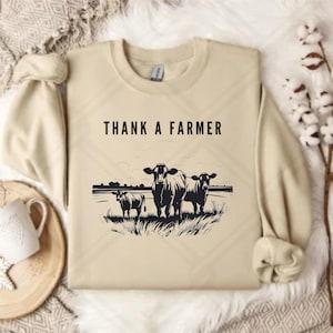 May include: Beige sweatshirt with the text "Thank a Farmer" printed in black. The sweatshirt features a black silhouette of three cows in a field.