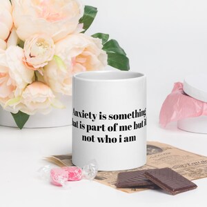 Könnte beinhalten: Weiße Keramik-Tasse mit dem Text "Anxiety is something that is part of me but it is not who I am" in Schwarz. Die Tasse ist von rosa Blumen, Pralinen und verpackten Süßigkeiten umgeben.